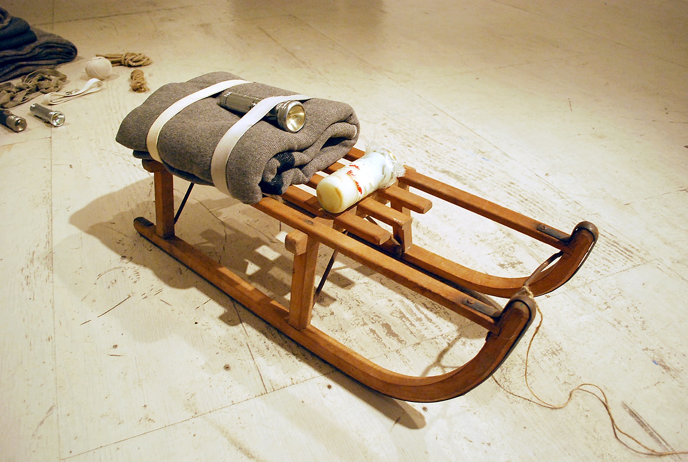 A small, old-fashioned, wooden sled with a gray folded blanket and silver flashlight strapped to the back with two white straps.