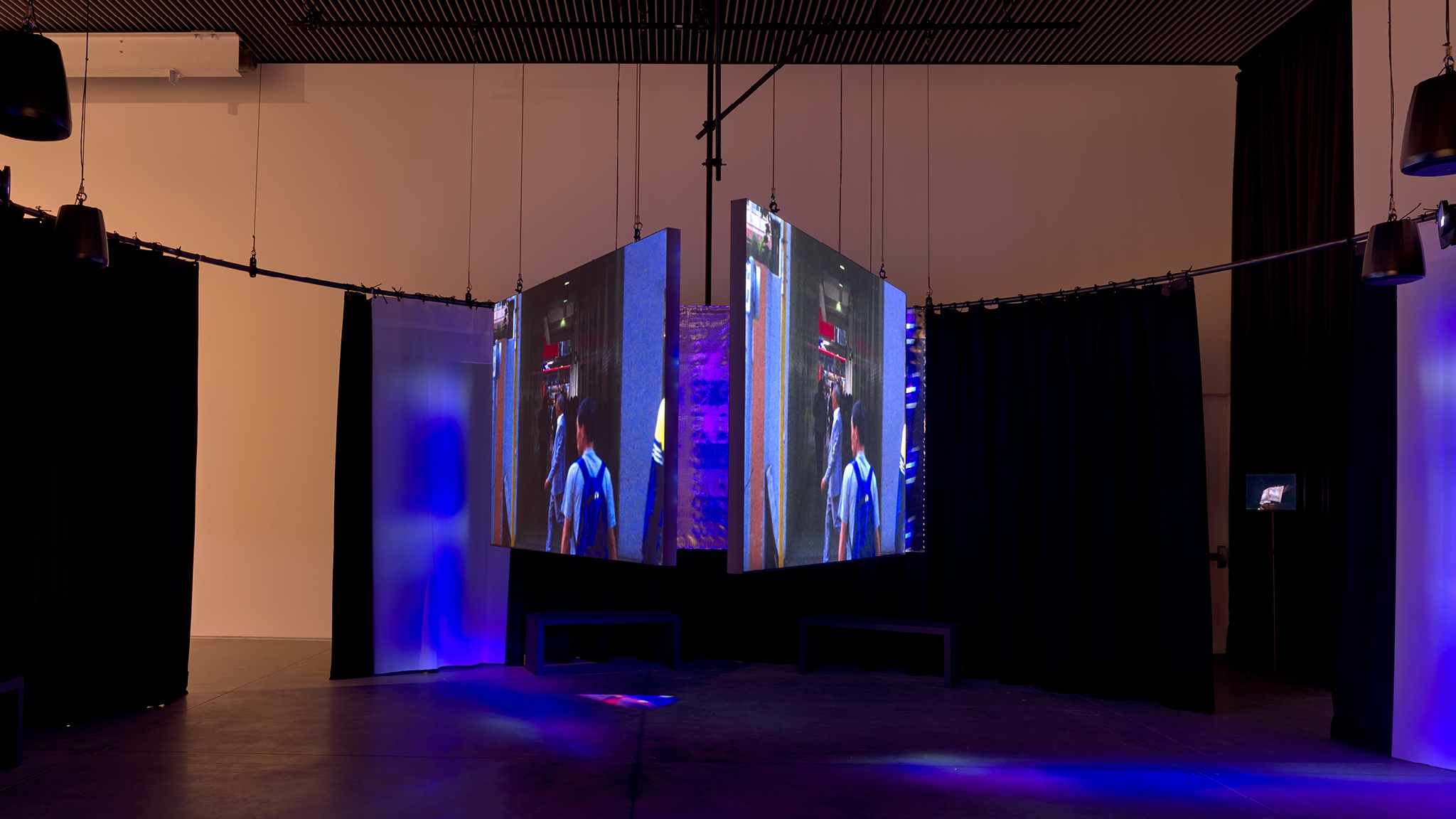 Three video screens hung in a triangular formation in a darkened space awash in bluish light. The screens depict people moving through a cityscape.