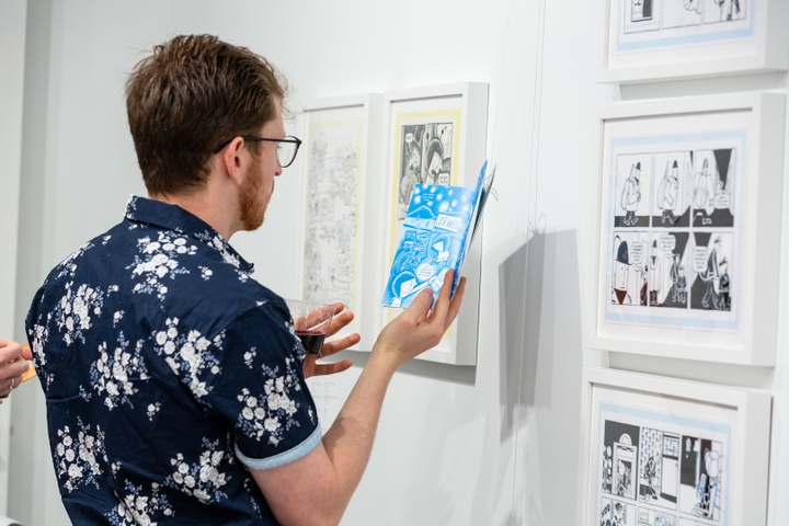 A visitor holds up a book in front of a wall displaying multiple black and white illustrations