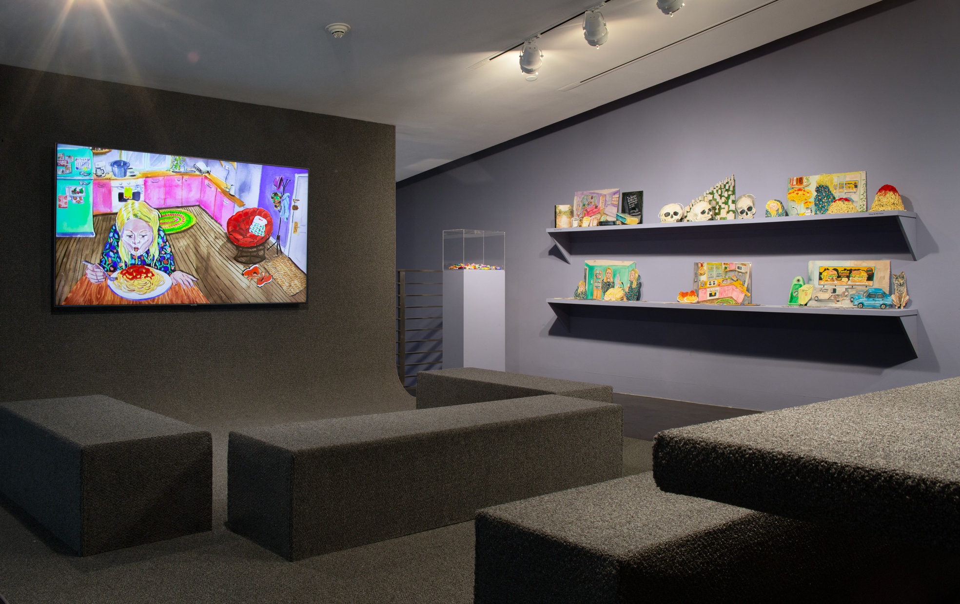 A gallery features gray carpeted floor and seating with a video animation playing on one wall and shelves displaying painted paper artworks on the other.