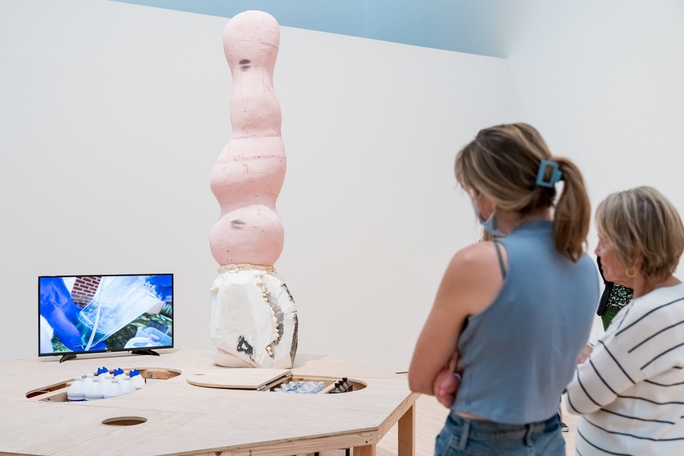 Two people watch a video of rubber gloved hands mixing chemicals. The tv is set on an unfinished, wooden stage at waist-height. A tall, rounded sculpture carved from pink foam sits next to the tv.
