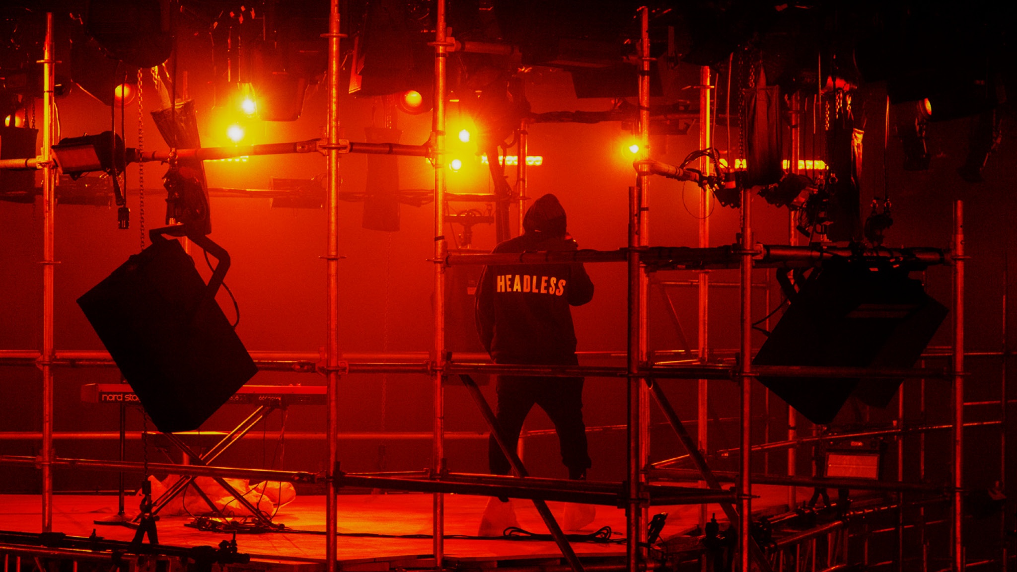 A performer wearing a hoodie and seen from behind stands on scaffolding in a theater lit by red spotlights