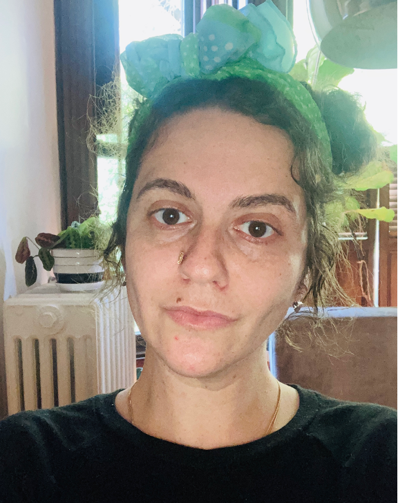 A photo of Rachel Katwan, who wears a green headband. A plant sits on a radiator in the background and light comes in through a window. 