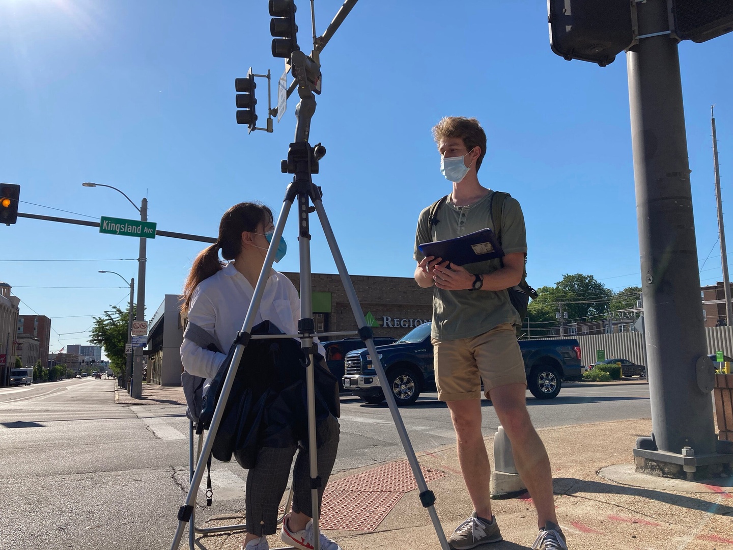 Two students at the intersection of streets, with a street sign for Kingland on a stoplight overhead. One student, seated behind a tripod, is speaking to the other student who is standing. Both are wearing face masks.