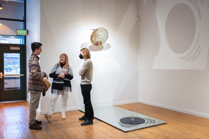 Three people stand next to a sculptural installation in a gallery involving reflections from a large sheet of mirror on the floor.