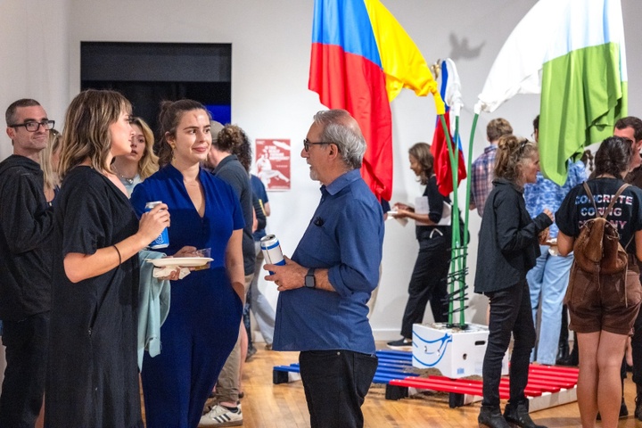Two students chat with Dean Colangelo in a gallery space. Behind them is a freestanding sculpture with three brightly colored flags hanging from bent poles.