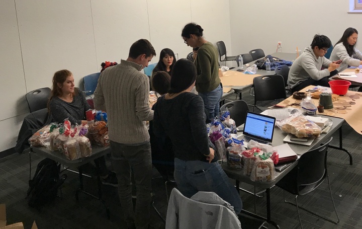 Students stand around tables with bags of bread on them. 