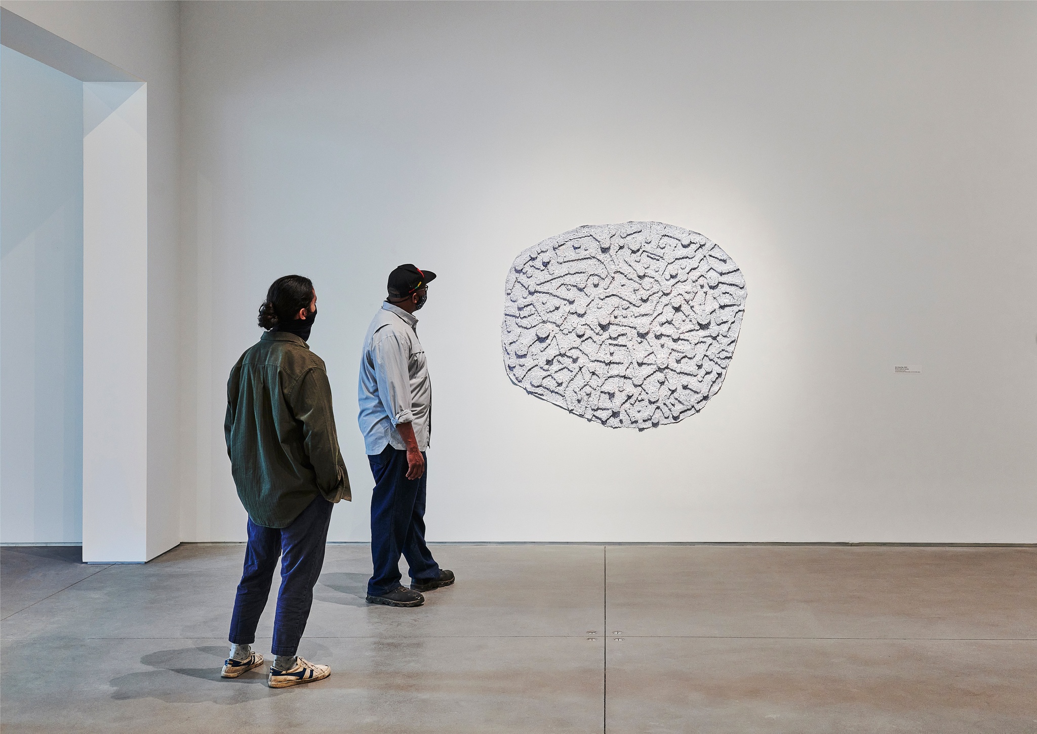 Two gallery visitors standing staggered from one another look at a curvilinear abstract painting in the exhibition Howardena Pindell: Rope/Fire/Water. The painting is a pale purple and its surface is covered in glitter, paper dots, and geometric forms that give it texture and depth.