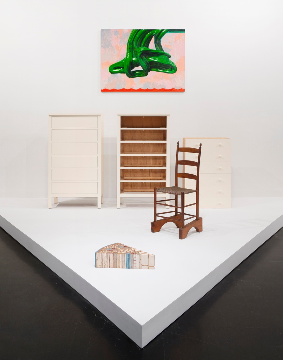 A pink and green artwork hangs on a white wall and below are three white dressers and a wooden chair with an abstract triangular object in front.