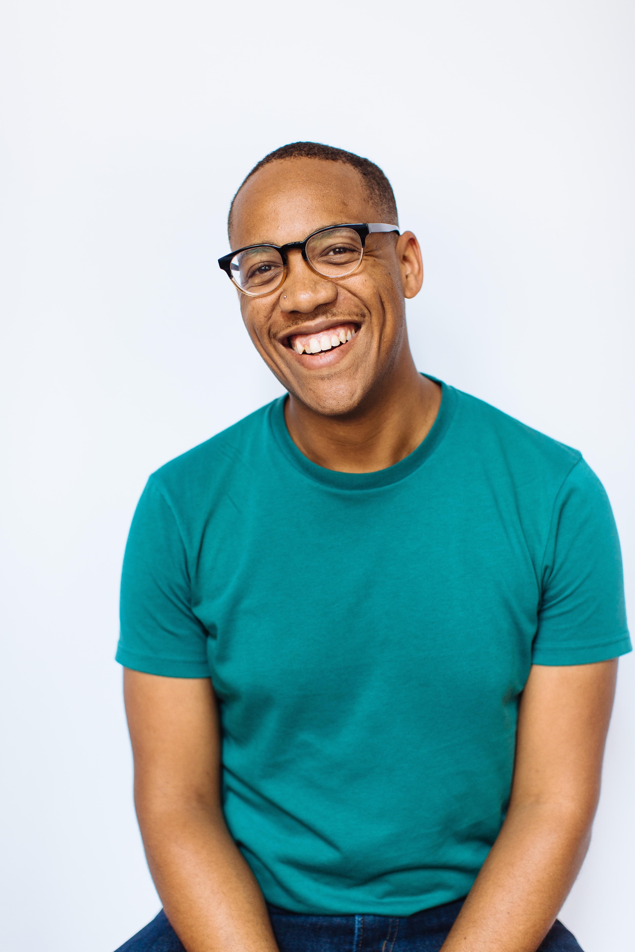 Artist Troy Anthony sits against a white background with hands in his lap, smiling broadly. Troy is a Black person with a shaved head who wears glasses and a teal t-shirt in this photo.
