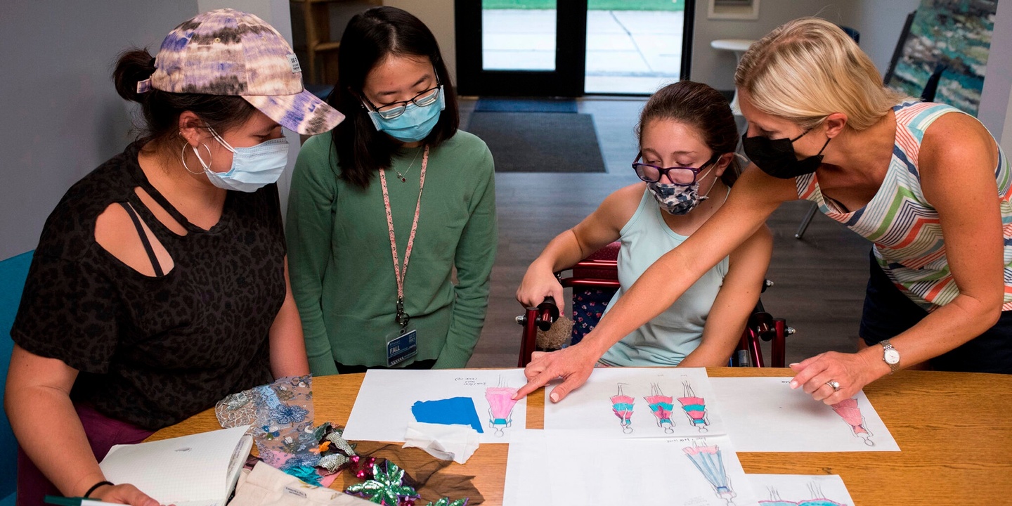 Four people wearing COVID masks gather around a table, looking at sketches of clothing designs.