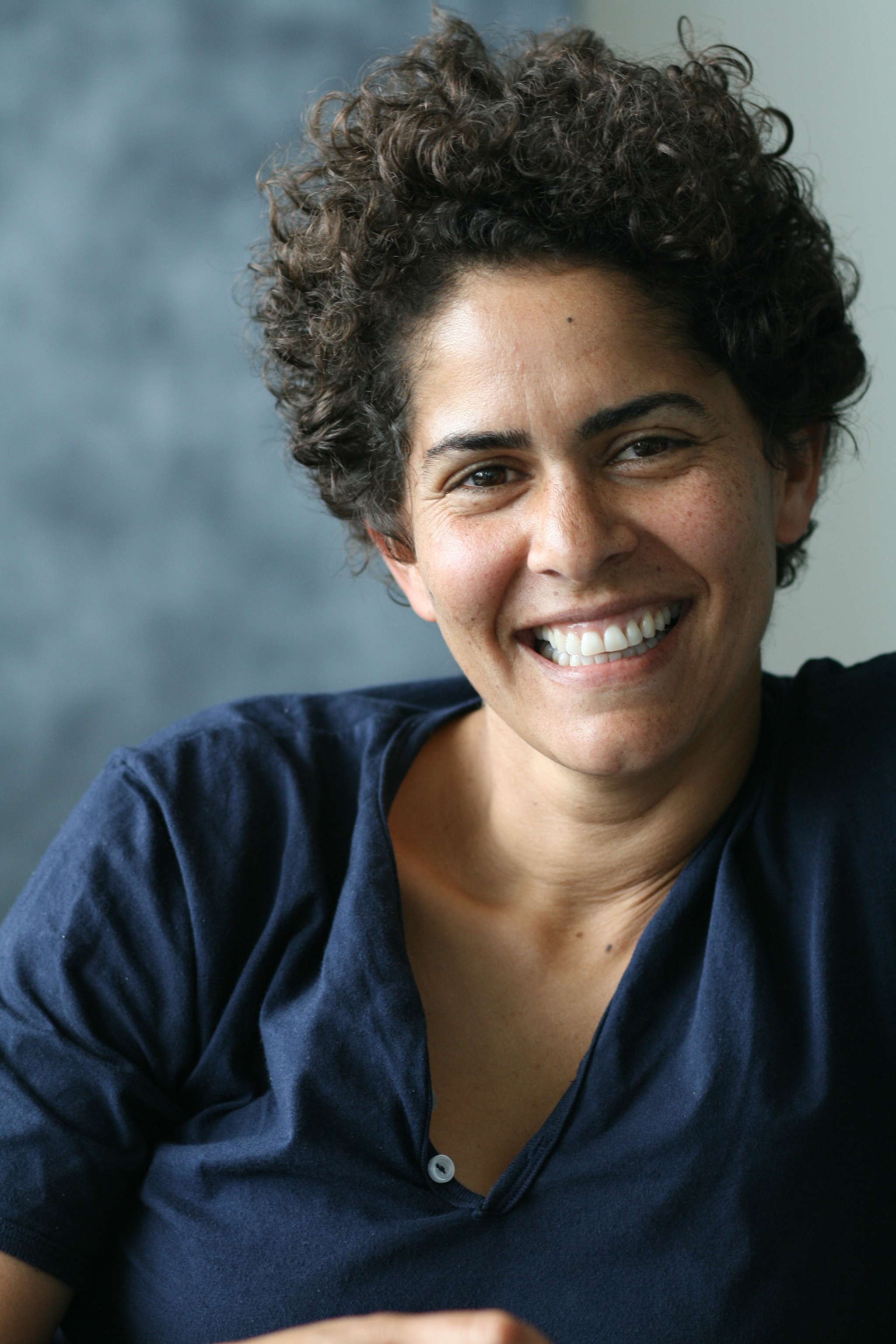 A photo of the artist Julie Mehretu smiling against a blurry gray and white background. Mehretu wears a blue shirt and light falls on the right side of her face.