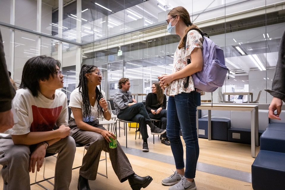 Hillawi Abraham sits in a chair next to students in a brightly-lit common room. One student approaches to ask a question.