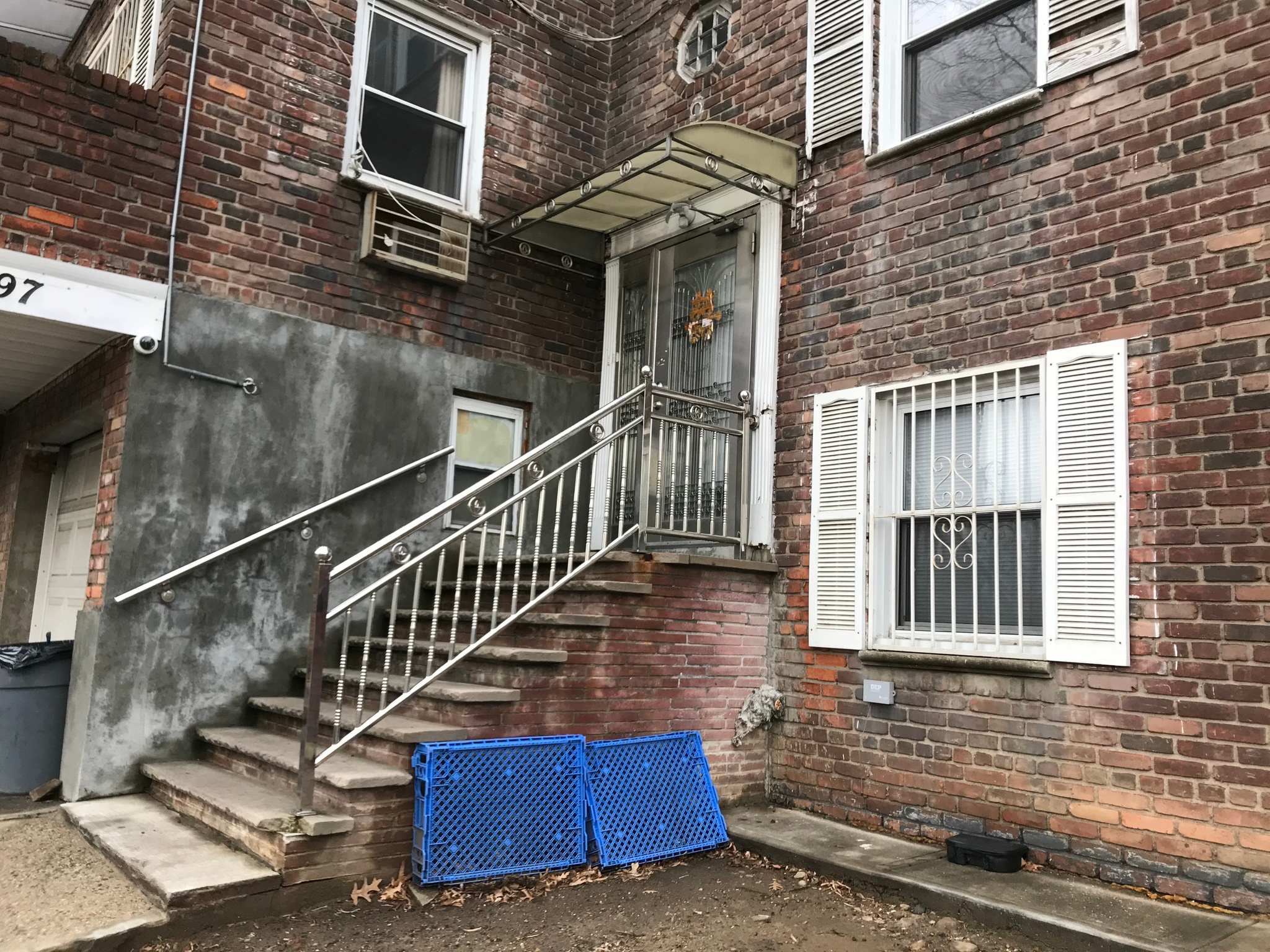 A stairway leading to a door of a house in a corner formed by two brick buildings. The door is made of steel, with a glass and metal awning above it, and a steel railing lining the side of the stairway. Against the stairway, two blue plastic containers are propped against the brick. 