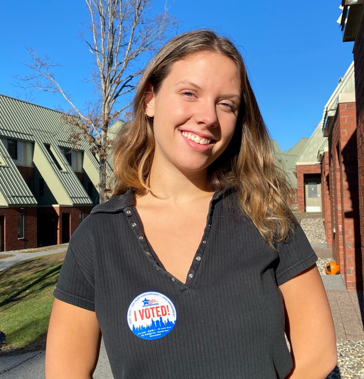 A young woman wearing a black shirt with a sticker on her chest that says, “I VOTED!” in bold red font smiles at the camera while standing outside in front of the Skidmore apartments.