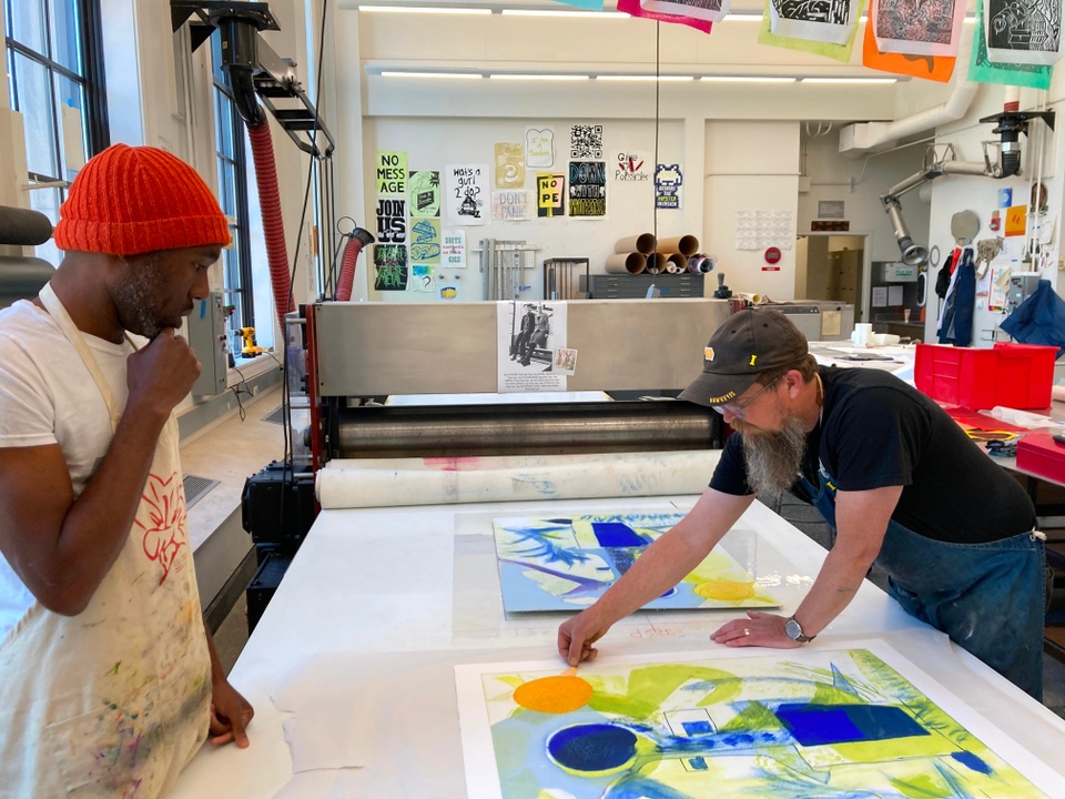 Tom Reed, master printer, leans over the print that was just pulled off the press while artist, Barber, watches with his hand on his chin.