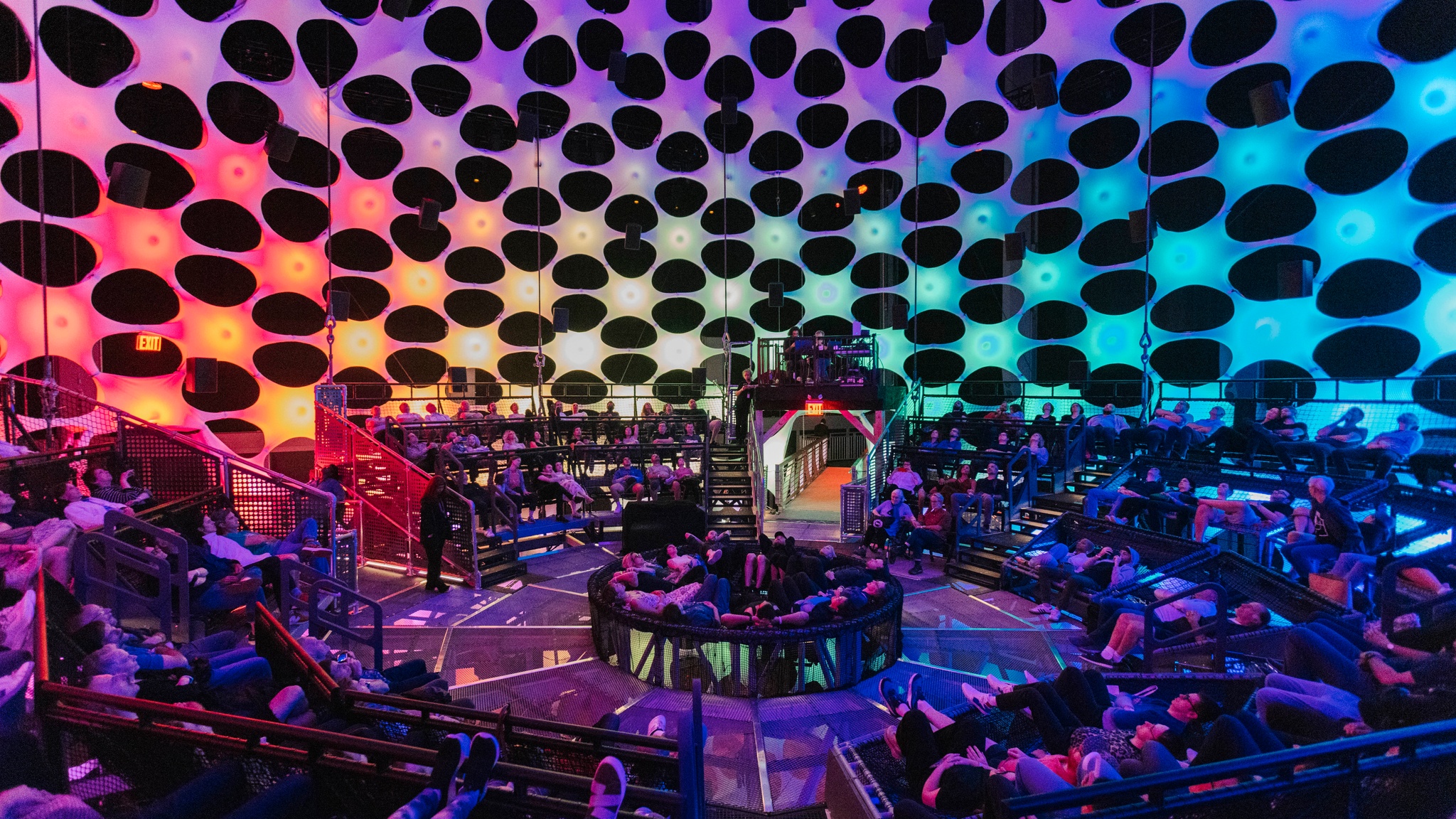 People in relaxed poses lie on their backs on netted seating banks inside a concert hall with a spherical wall. The wall is porous and composed of light nodes that emanate orange, purple, and blue light.