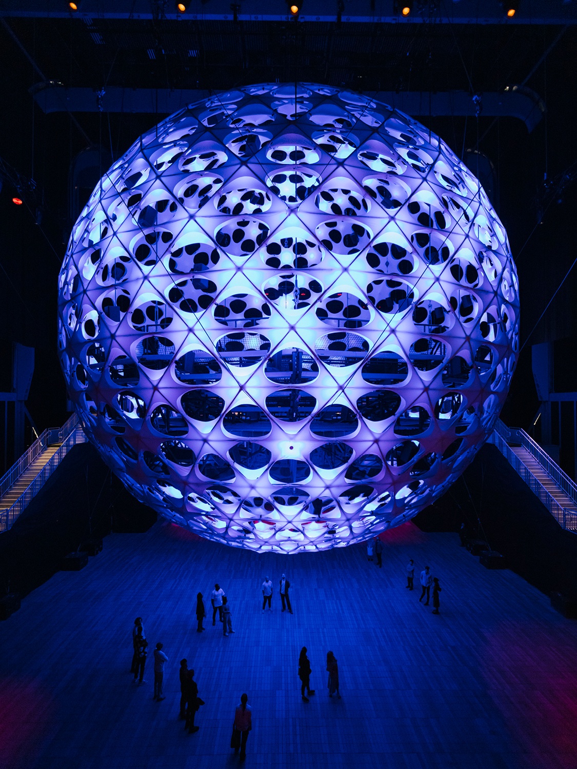 A large spherical structure hangs from a 115-foot-tall ceiling above roughly 20 people who stand below it. The sphere is dotted with light nodes that emanate a purplish blue light. The people are small figures beneath the enormous sphere.