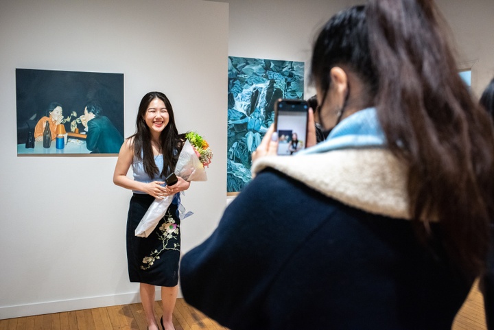 Person holds flowers and stands in front of a painting of a man and a woman sitting across from each other at a table in a dark room, drinking something out of a tall glass bottle.