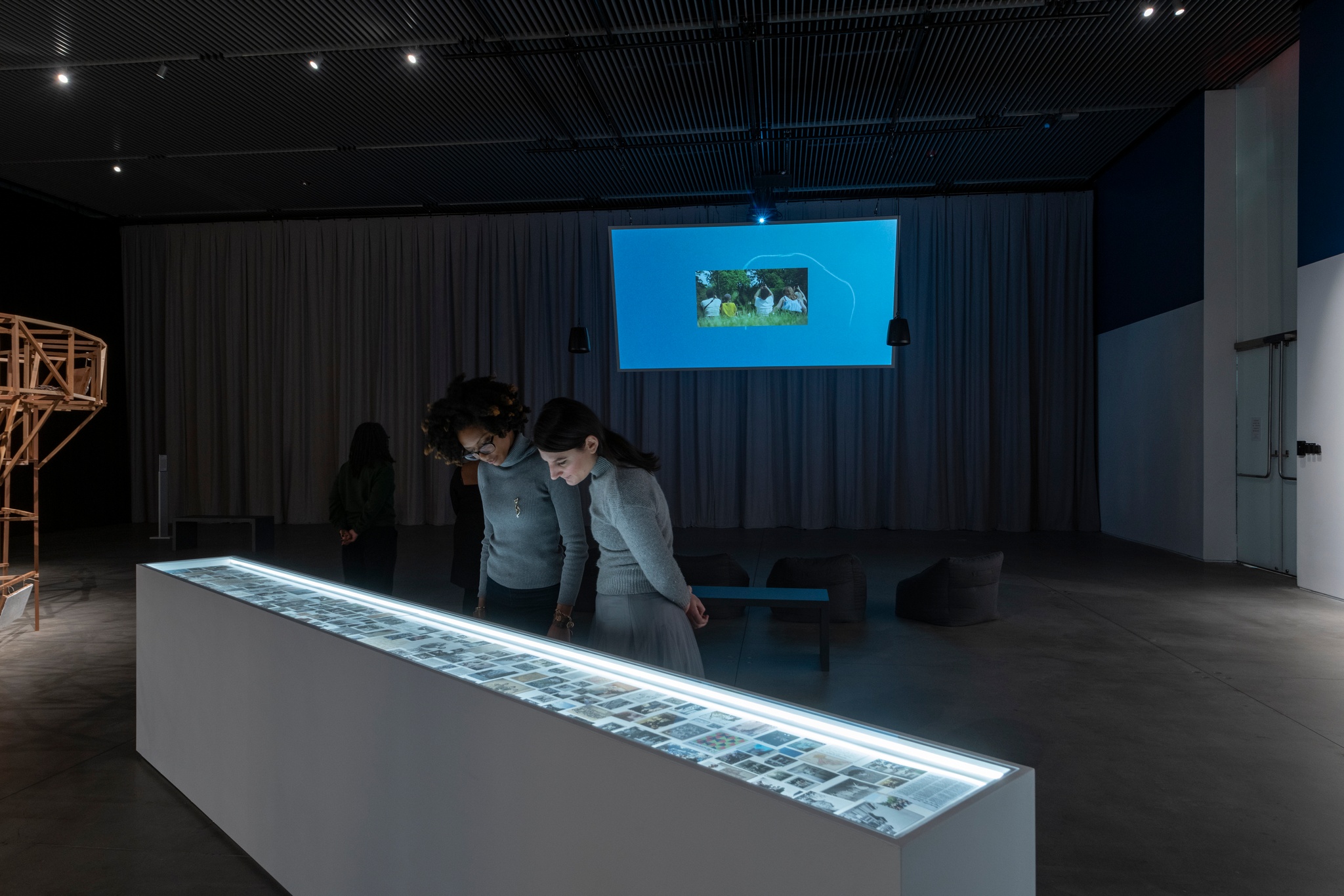 Two gallery visitors peer into a long, lit vitrine. They are examining images as the lights from the vitrine illuminate their faces. Behind them, the gallery is in shadow and a film screen hangs from the ceiling. The screen shows an image of a blue sky. 