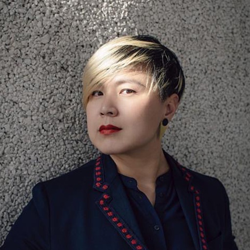A Chinese woman with short hair bleached in the front along her bangs looks at the camera with one eye highlighted by a stripe of sunlight shining on her and the gray cement wall behind her. 