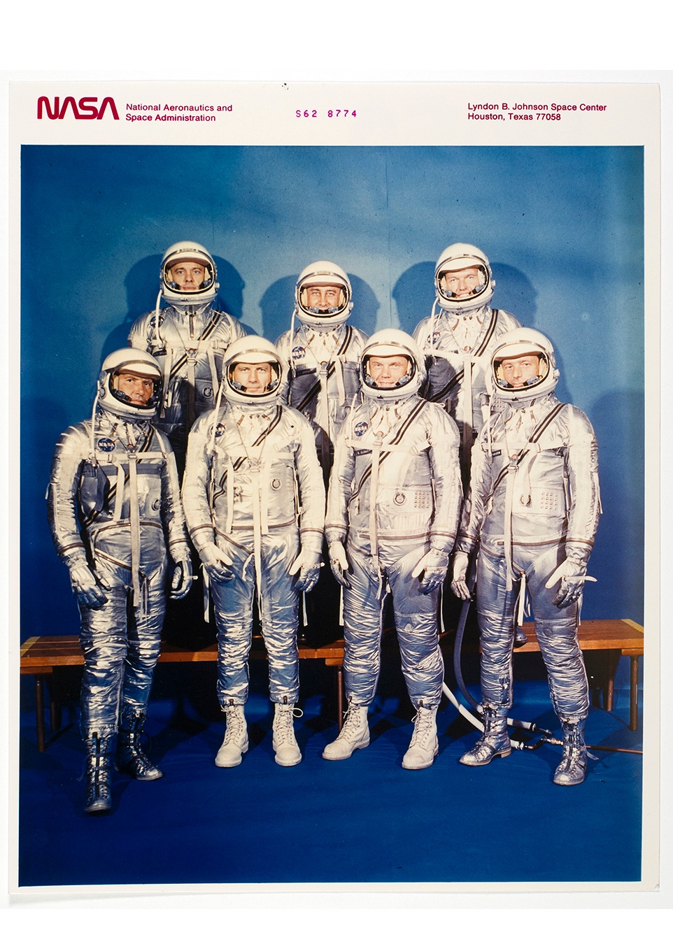A group photograph of seven men in shiny silver space suits pose for a portrait in front of a blue background.