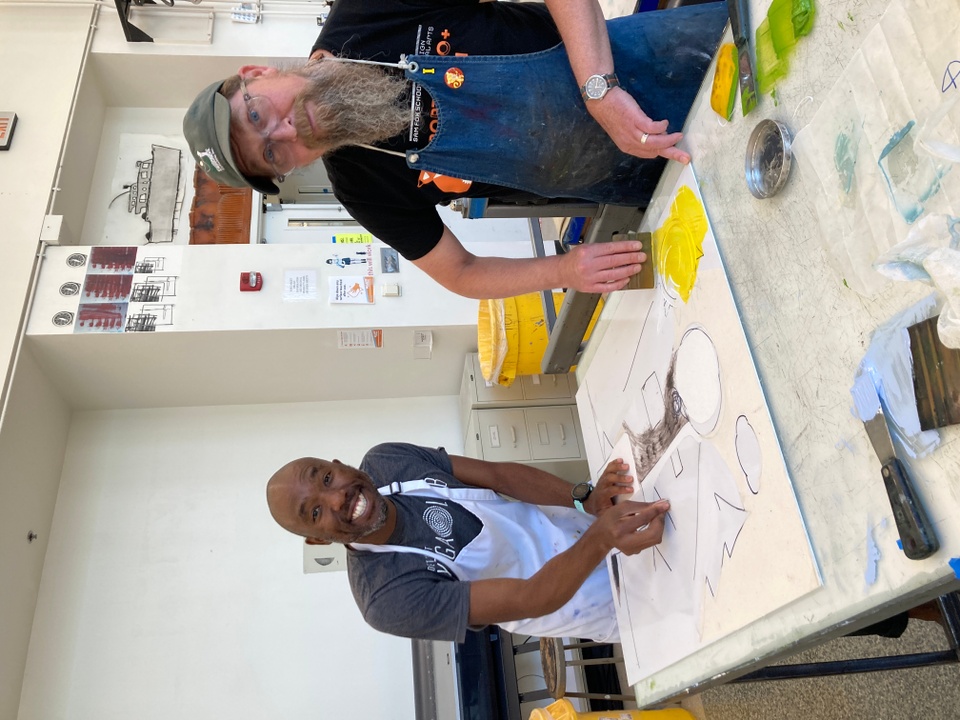 Two people, the artist bARBER and the printer Tom Reed, stand next to a glass table in preparation for inking a plate and printing.