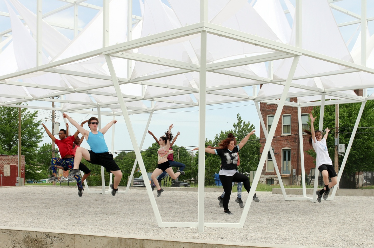 A group of people leap in the air under an elevated white structure with sails. 