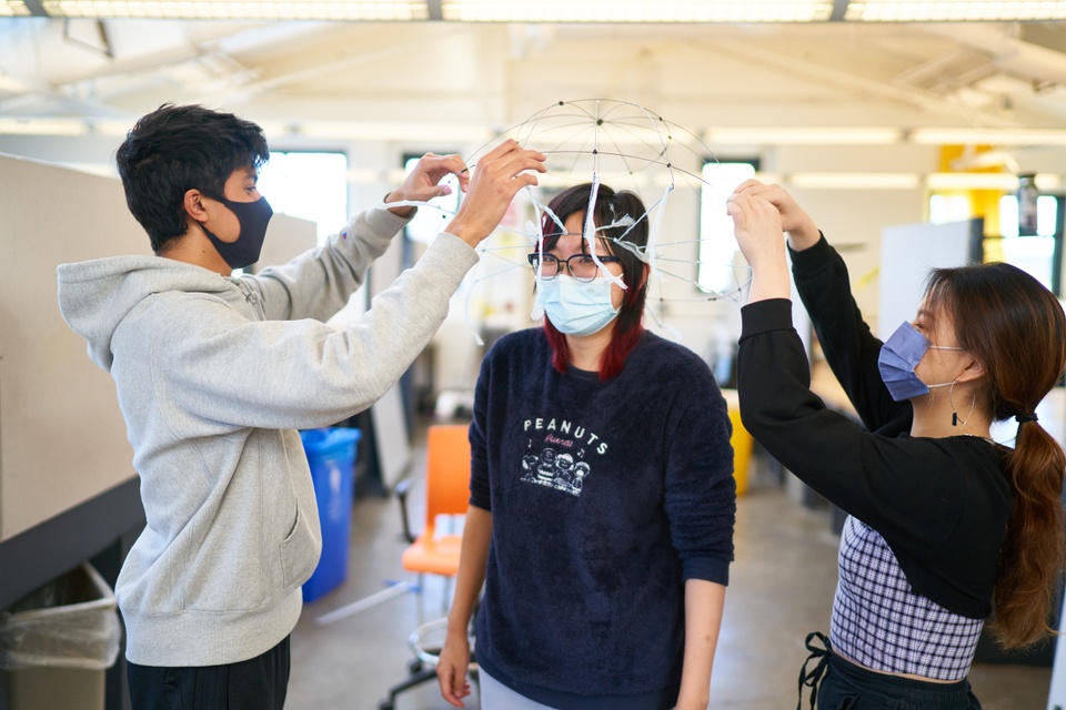 Three students in a studio space, with two standing to the sides of the one in the middle, arranging a wire, domed hat structure over their head.