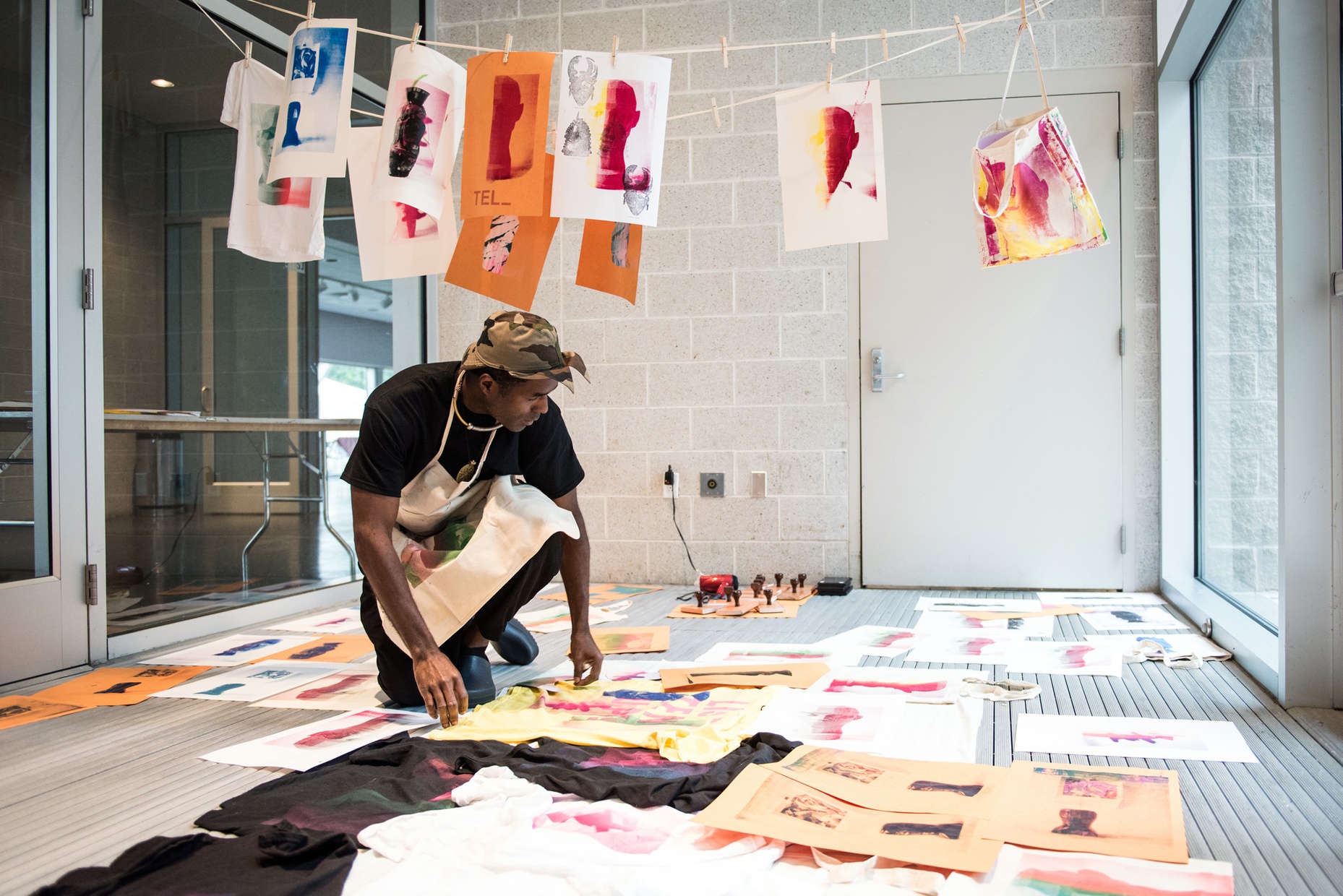 A Black man in an apron crouches in a space filled with prints on paper and t-shirts laid out to dry.