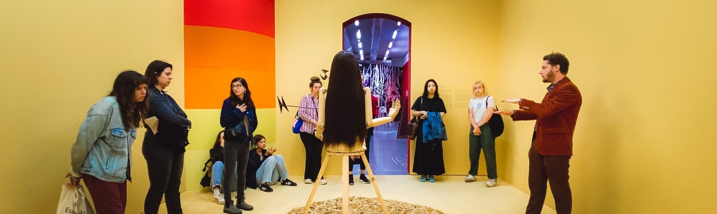 A group of people stand in a circle looking at a sculpture while one person leads the discussion.
