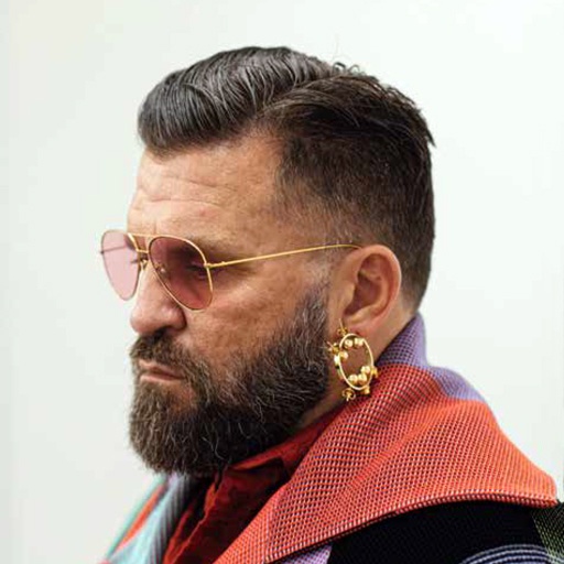 A man seen in profile wearing metal-rimmed aviator-style glasses and a gold hoop earring with a dark beard and combed hair cut in a fade on the sides of his head. 