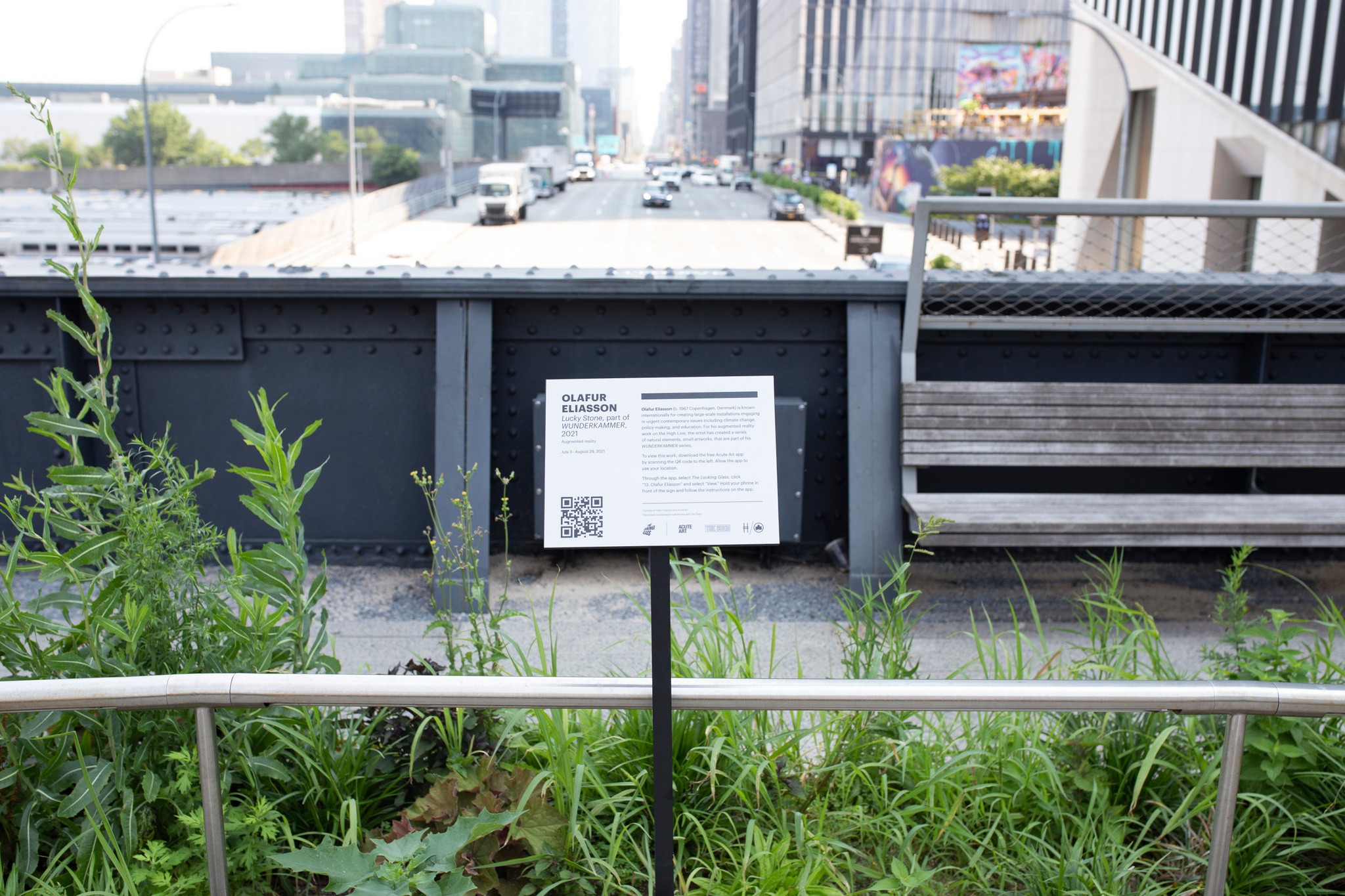 An artwork label on a signpost installed along wild grasses on an overpass on the High Line with a street passing beneath and stretching into the distance
