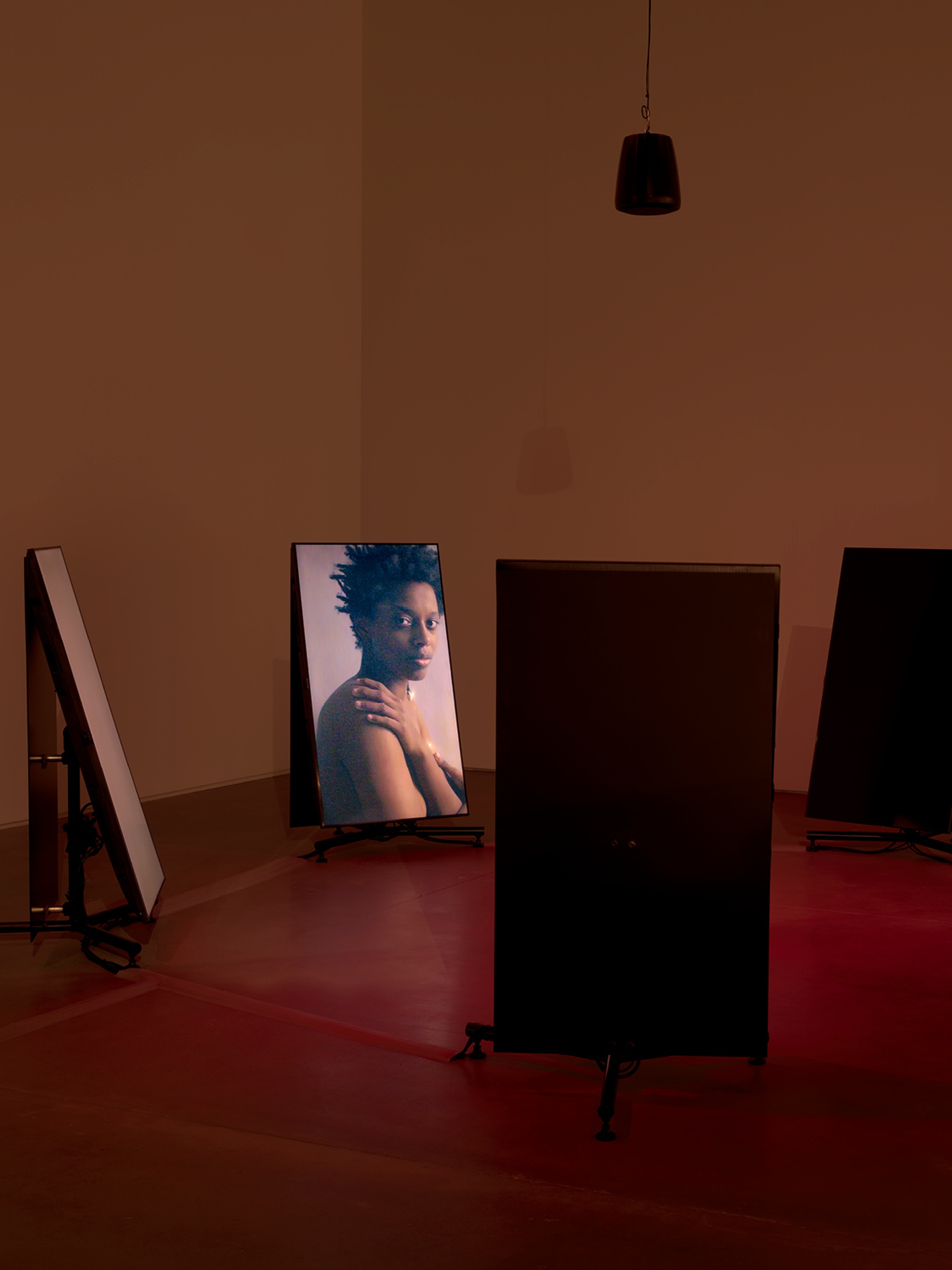 Four vertical video screens stand in a circle in a space cast in an ambient red light. On one screen facing the camera, the artist Le'Andra LeSeur is depicted with bare shoulders turning her head to look out at the audience. 