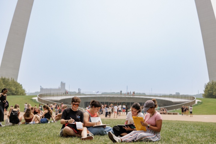 Four students sit on the grass under the St. Louis Gateway Arch and sketch.