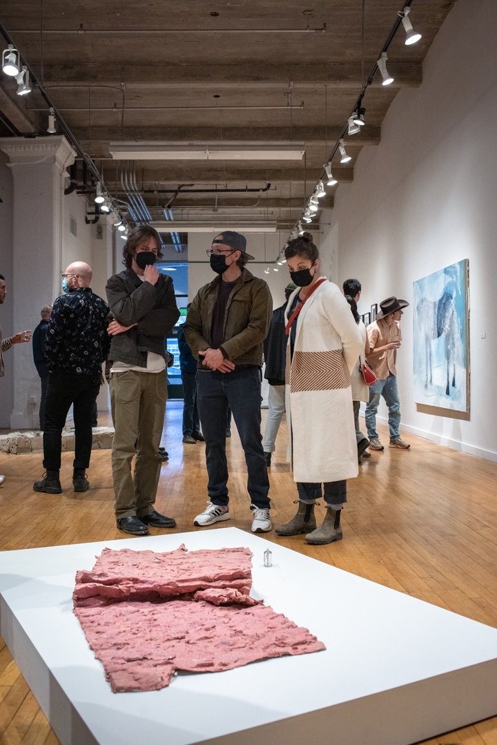 People stand around a short white platform on a gallery floor, on which rests a crumpled pink roll of paper or fabric.