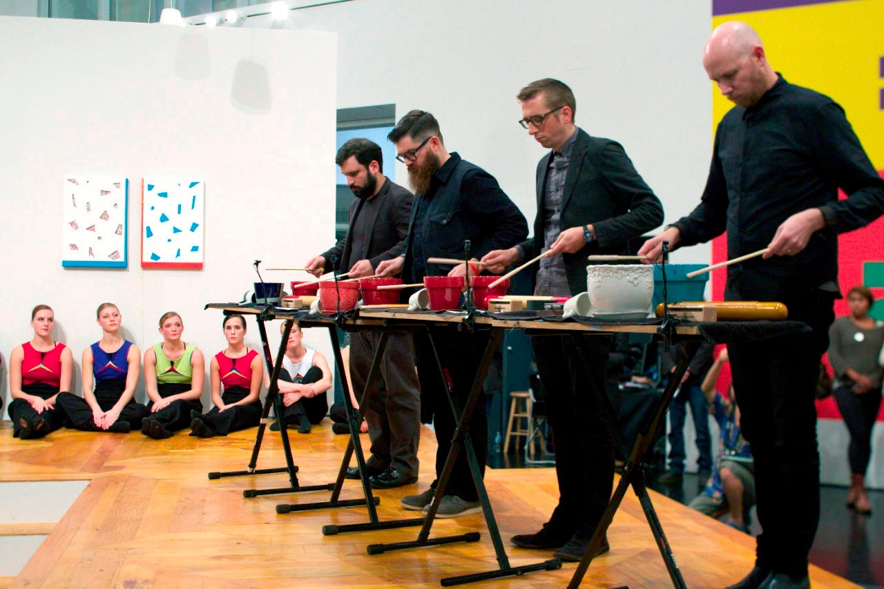 A group of four, light-skinned men stand in a row on a wooden stage using wooden sticks to beat on different ceramic bowls and mugs while a group of people look on in the background.
