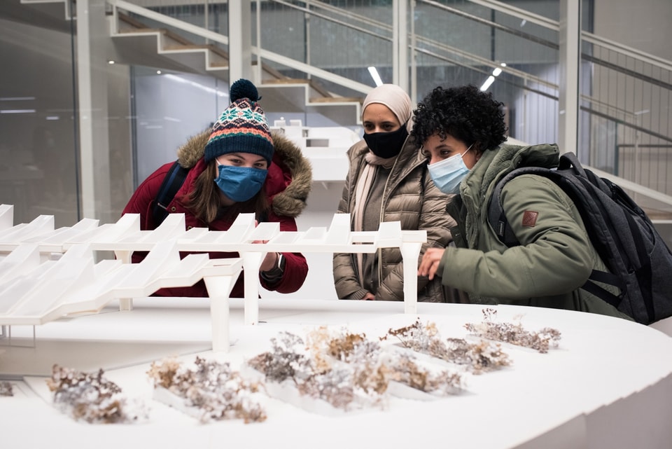 Three people bend down to look inside a table-sized white architectural model of a pavilion surrounded by model shrubbery.