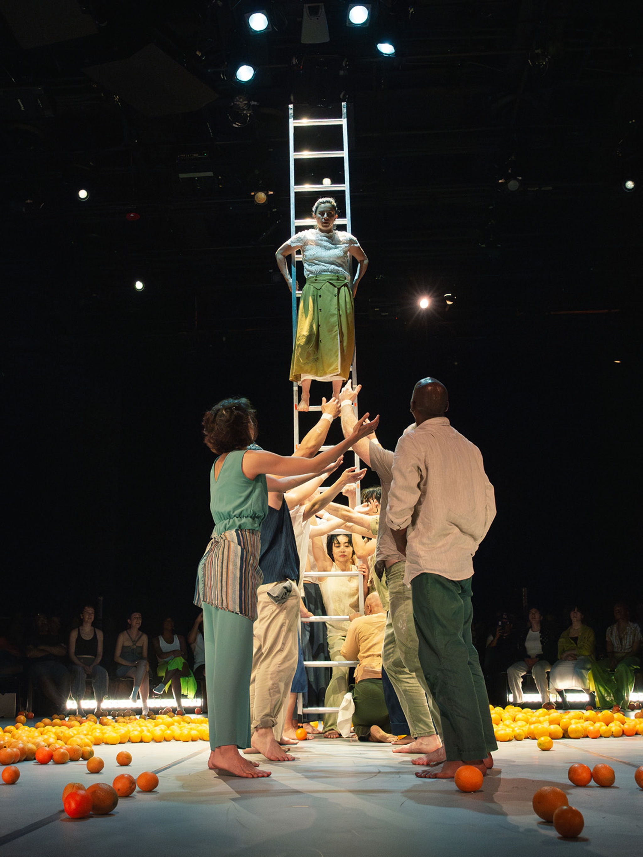 A group of dancers holds a metal ladder erect in the center of a circular performance stage. One dancer has climbed the ladder above the others' heads. Around them are strewn brightly colored oranges. 