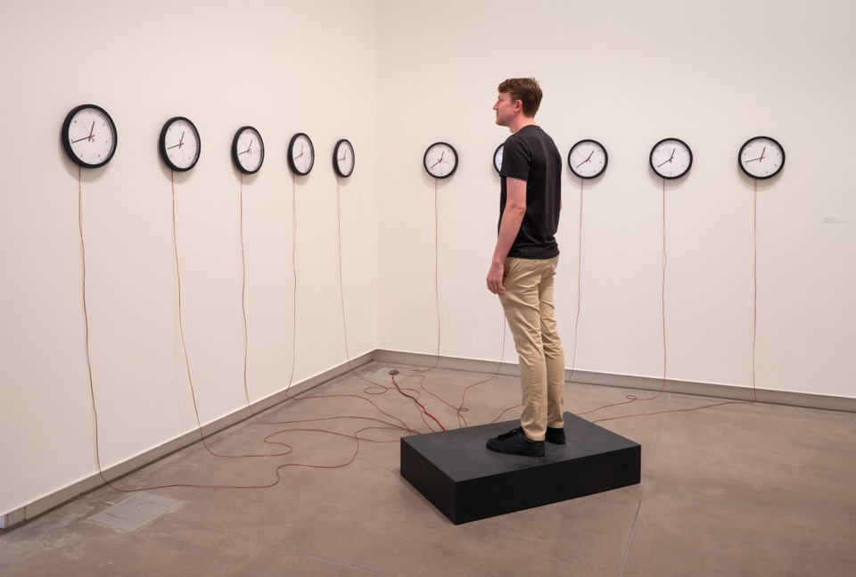A person stands on a black wooden box and views a row of ten analog wall clocks connected to the box with wires. Instead of numbers the clocks have an arrangement of letters.
