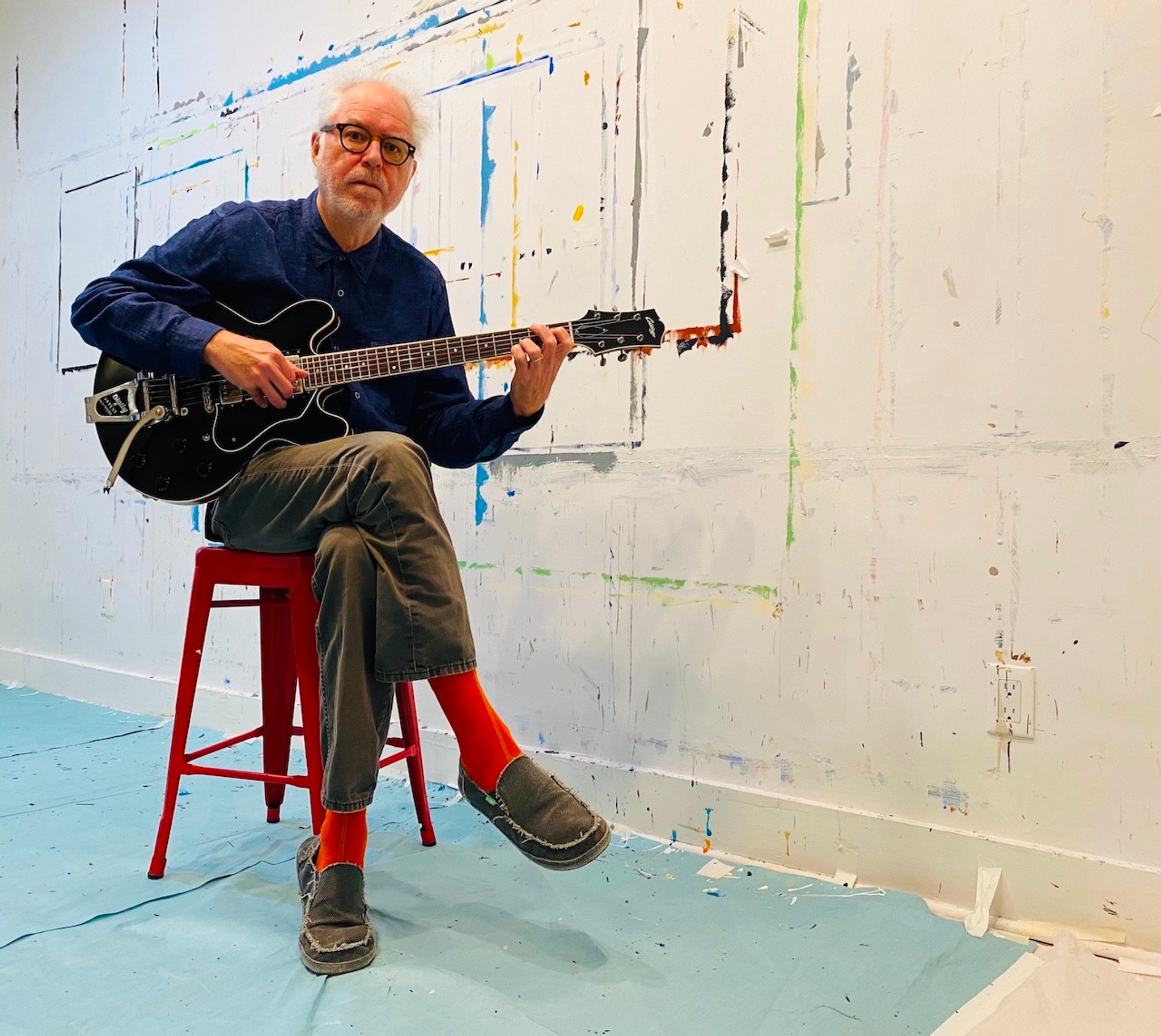 The guitarist Bill Frisell seated on a red stool with one leg crossed over the other where he balances his guitar. He is wearing bright red socks, brown pants, and a blue shirt and is sitting in front of a wall splattered with paint. 