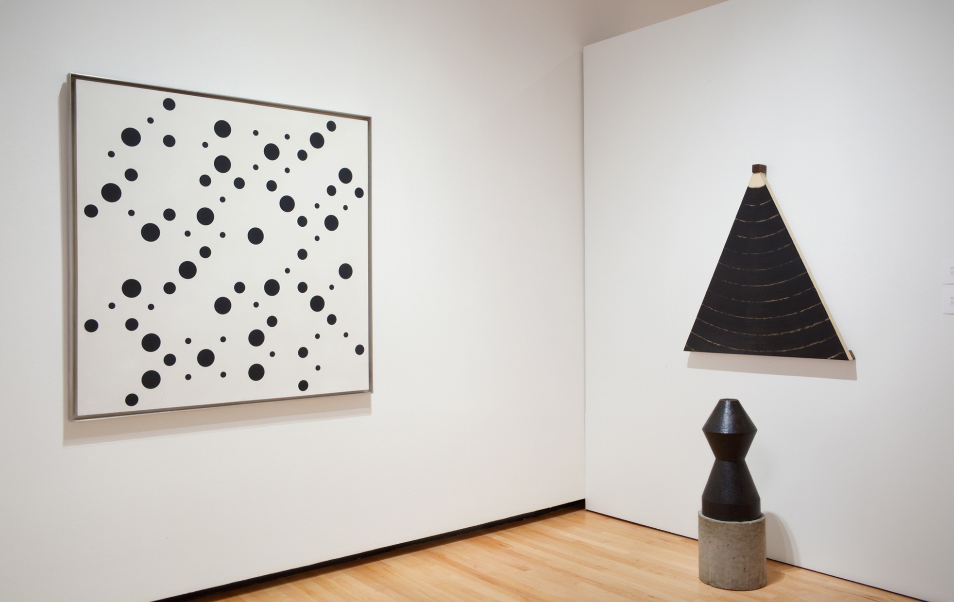 The corner of a room with a white painting with black dots hanging on one wall and a triangularly shaped painting hanging on the other wall with a sculpture below.