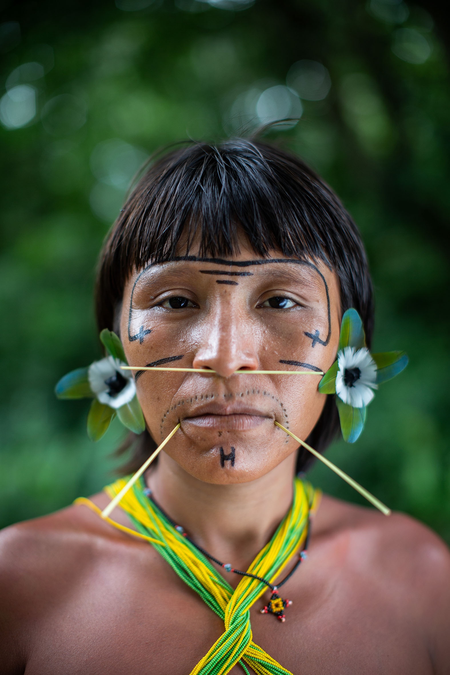 A portrait of artist Ehuana Yaira. An Indigenous Yanomami woman, Ehuana is seen from the shoulders up. She wears a garment whose green and yellow straps cross over her chest and around her neck. She has dark hair in straight bangs across her forehead. She has a long, straight piercing through the septum of her nose and wears a blue flower in her hair tucked behind either ear.