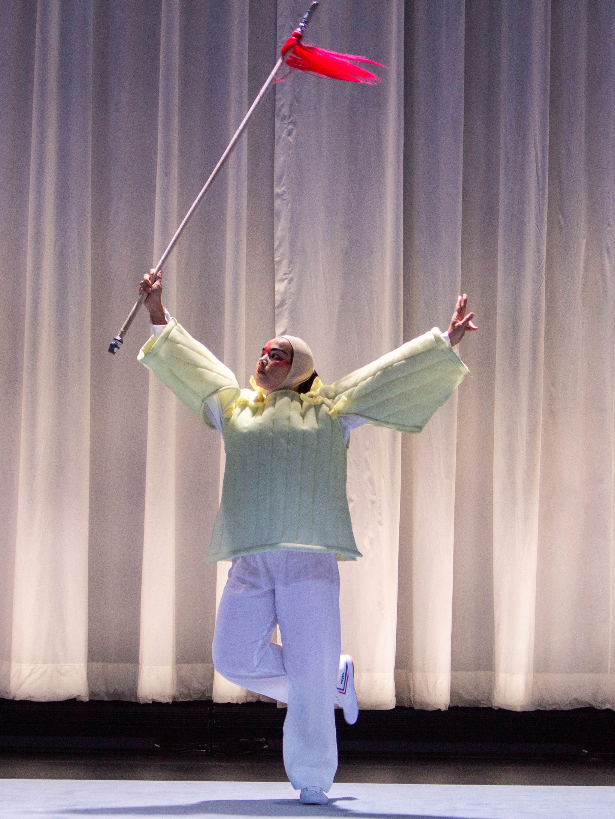 A dancer wearing a structured, loose fitting yellow top and baggy white pants stands with arms outstretched. She is looking upward and holding a pole-like staff above her head. The tip of the staff is tied with a small red flag.