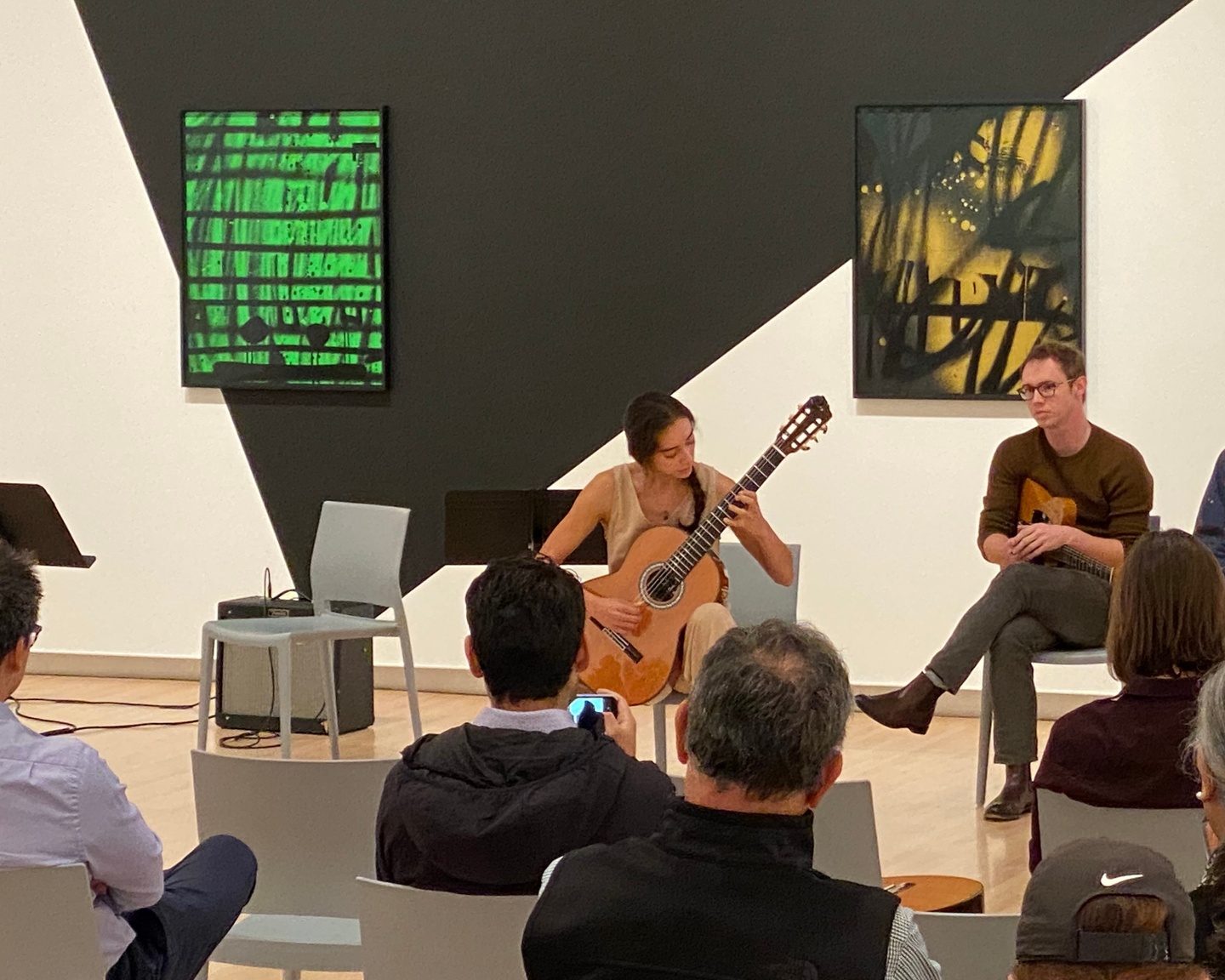 A person plays guitar in front of a wall with paintings and a black triangle painted on it, while other people look on