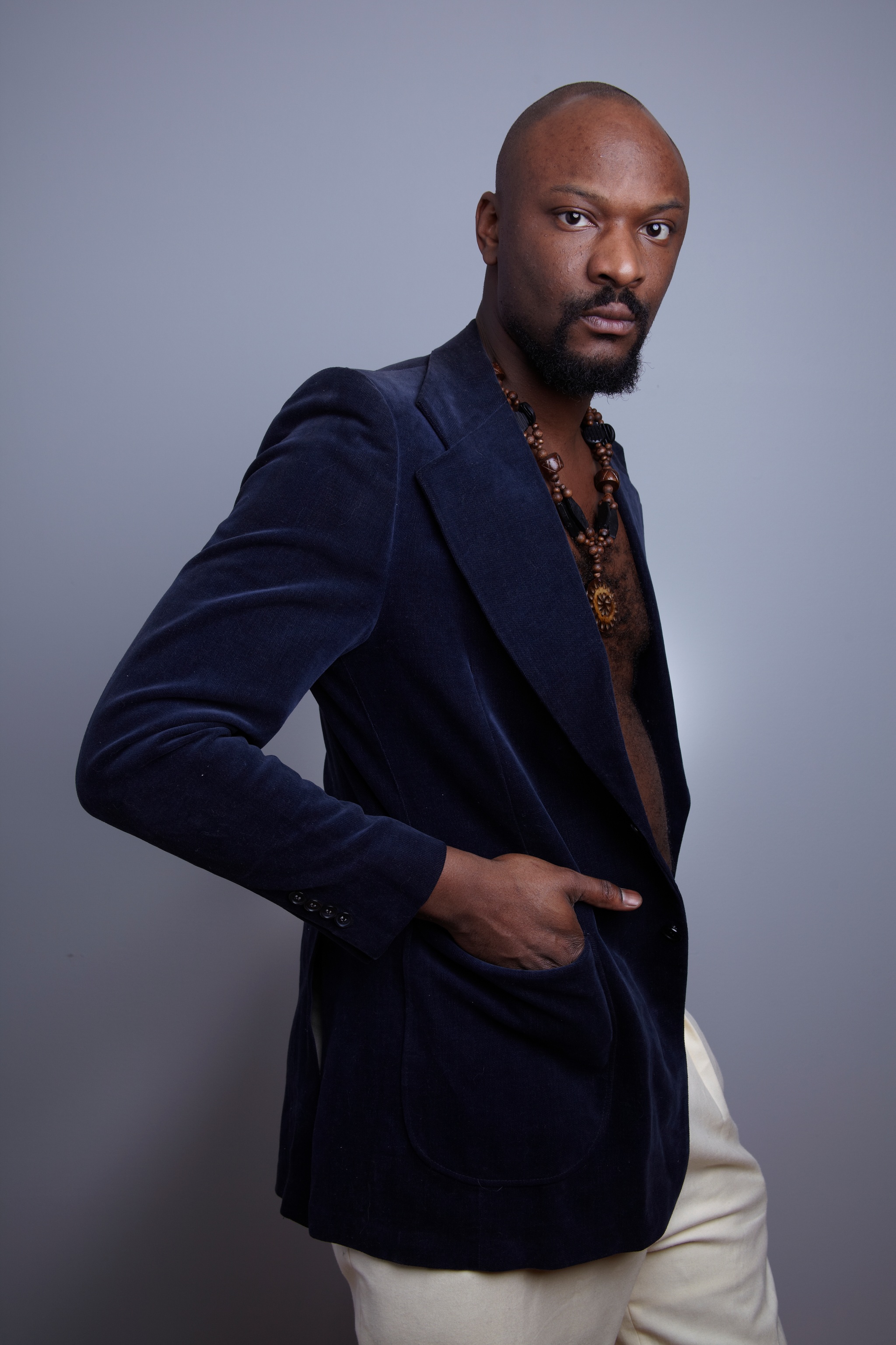 A portrait of the artist Justin Hicks standing against a light gray background in a navy blue blazer and white pants, with one had in his jacket pocket