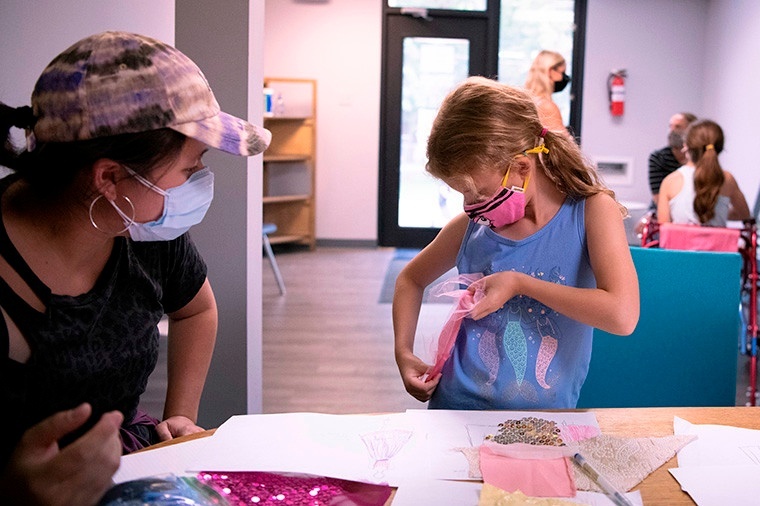 A younger child holds up a pink, gauzy piece of fabric to her waist while an older student designer watches. Both are wearing COVID masks and standing at a table with sketches on it.
