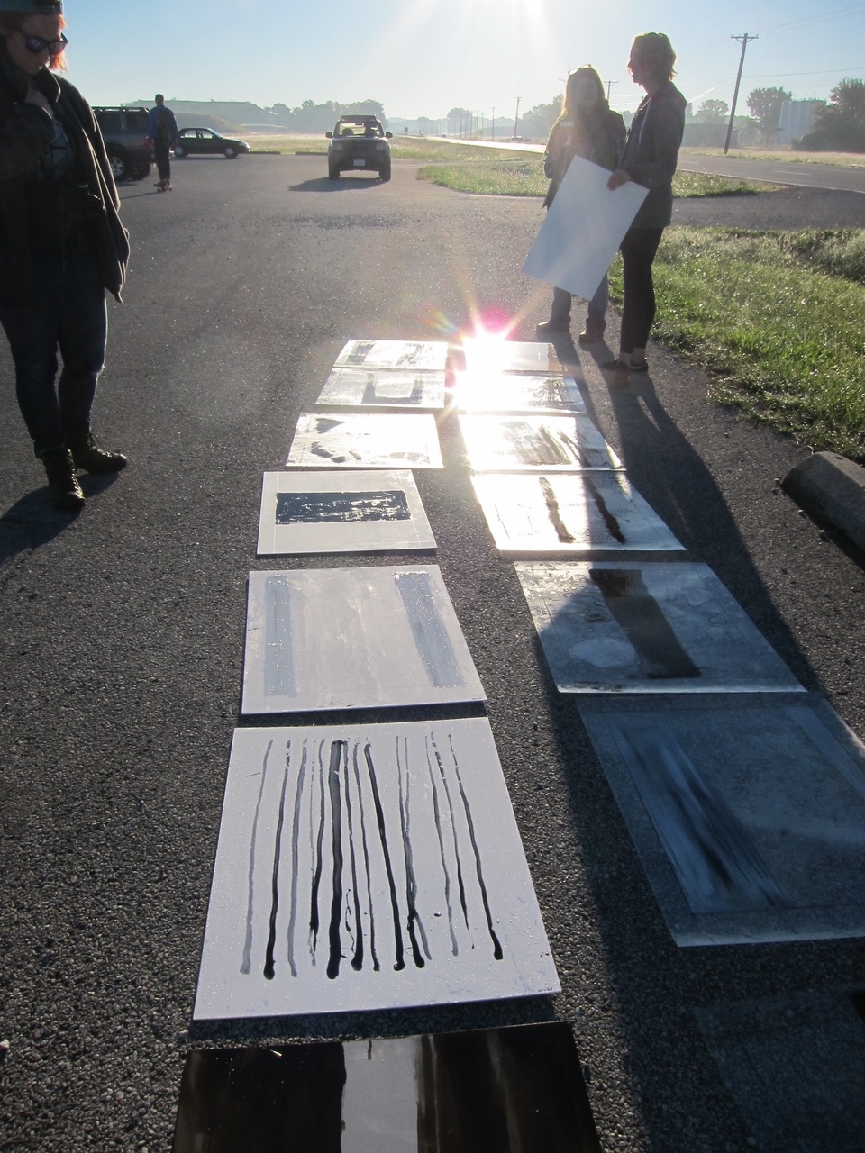 Various rectangular paintings with black and grey painted lines on them laid out on the roadside by a field