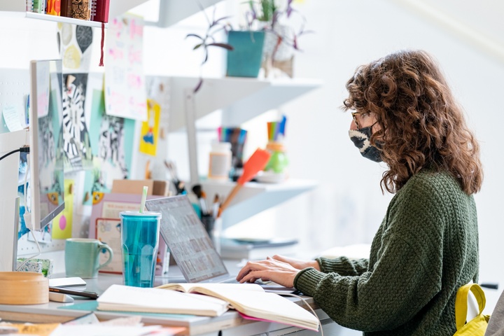 Person works on a laptop at a white desk decorated with many reference images and inspirational artwork, as well as knicknacks and a potted plant.