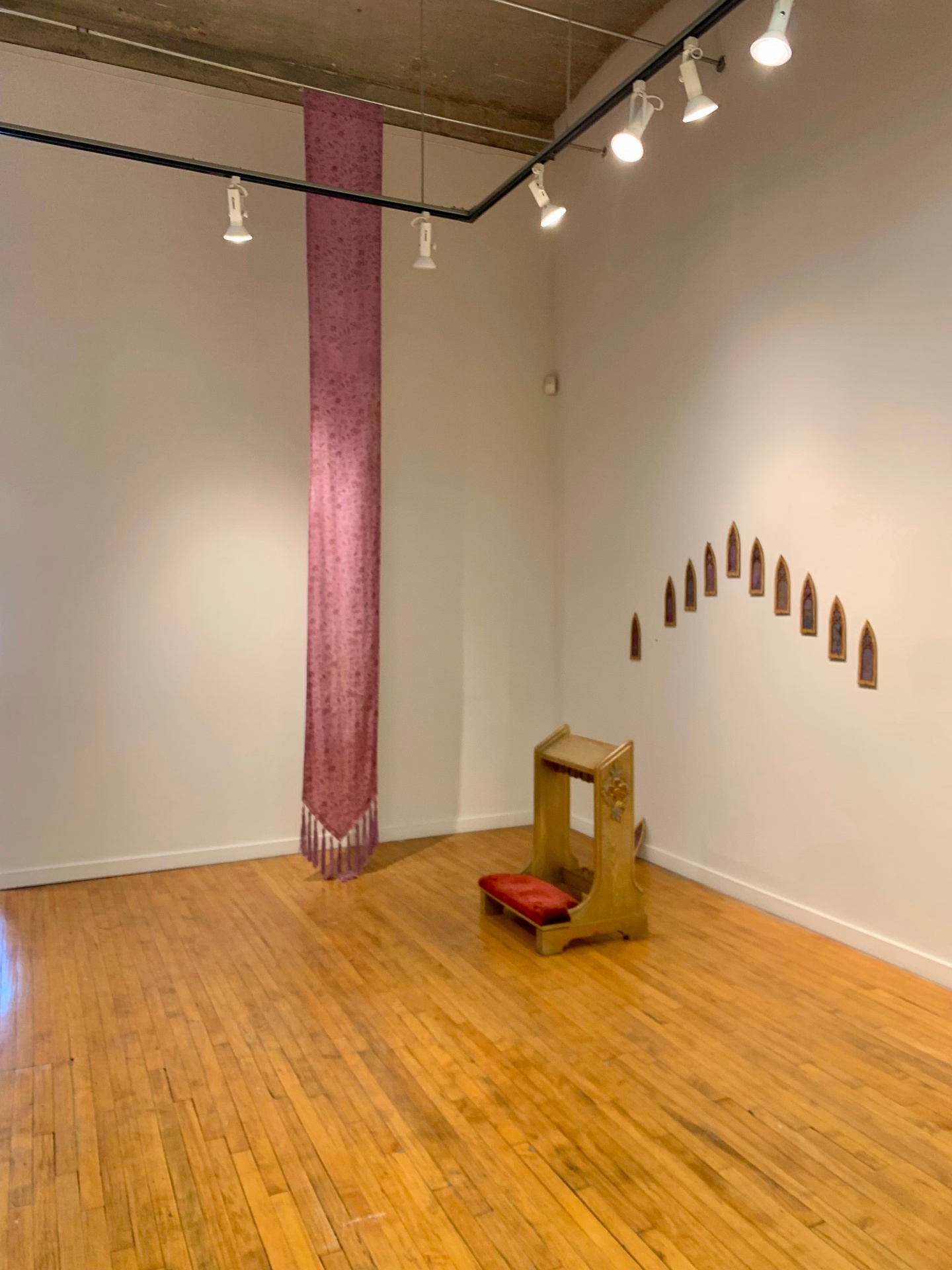 A church pew in front of several paintings resembling church windows with a pink tapestry beside it. 